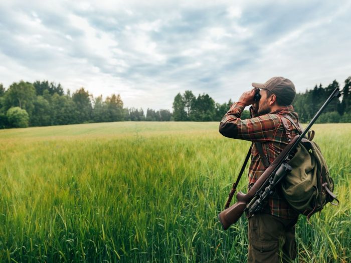 Mann mit Gewehr über der Schulter schaut mit Fernglas über ein Feld